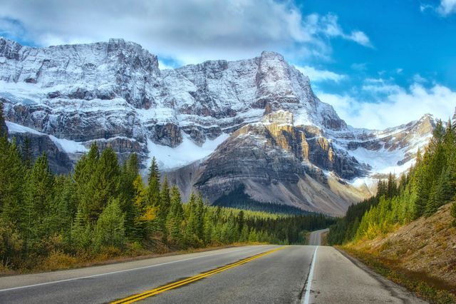 Amazing view of a highway stretching towards majestic snow-capped mountains under a blue sky in Banff National Park. Perfect for travel and adventure themes, advertisements for road trips, nature, and outdoor activities.