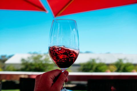 Hand holding swirling red wine glass under red umbrella on sunny terrace vineyard