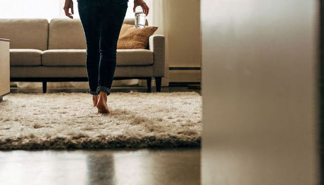 Barefoot woman walking on shag rug holding glass of water in sunlit living room