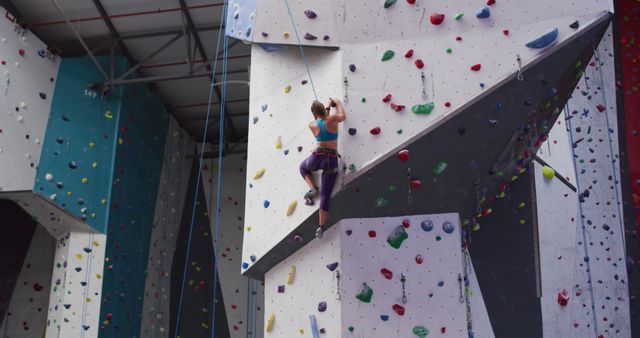 Woman climbing a vibrant indoor rock wall in a gym. Person is secured by a harness and ropes, emphasizing safety. Great for depicting fitness, athletic pursuits, rock climbing equipment, and indoor or gym activities. Useful for blogs, promotional materials, and inspirational fitness content.