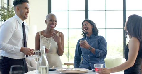 Laughing diverse professionals socializing in bright lounge with drinks and platters