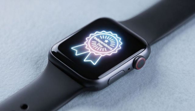 Dark gray smartwatch displaying neon independence day badge on soft pale blue surface