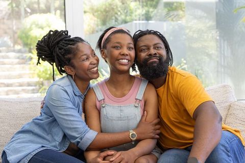 African american family smiling and embracing daughter on sofa at home with sunlight