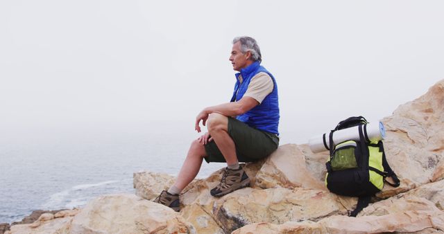 Senior hiker man with backpack sitting on the rocks in the mountains and watching the sea shore. trekking, hiking, nature, activity, exploration, adventure concept.