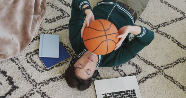 A young male teenager is relaxing at home, lying on the carpet holding a basketball, surrounded by a laptop and books. This can be used for depicting youth lifestyle, study breaks, balancing schoolwork and leisure activities, and relatable moments for teens.