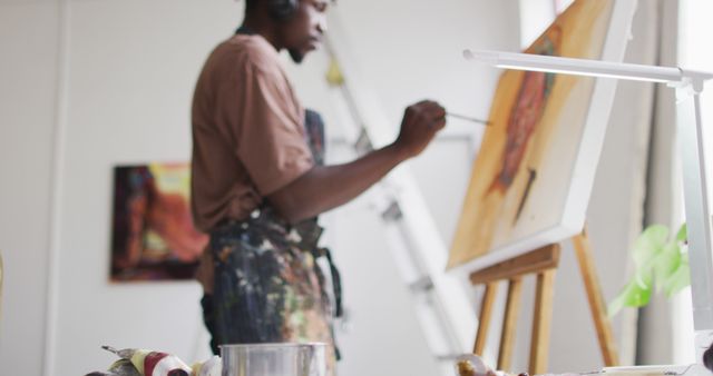 African american male artist wearing headphones painting on canvas at art studio. art, hobby and creative occupation concept