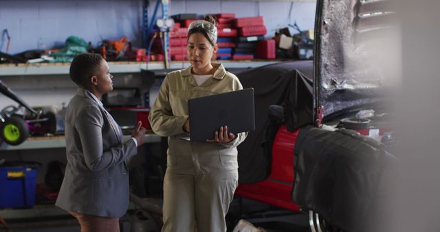 Image of two diverse car mechanic and business owner with laptop searching for car parts. working in car repair shop and running small feminine business concept.