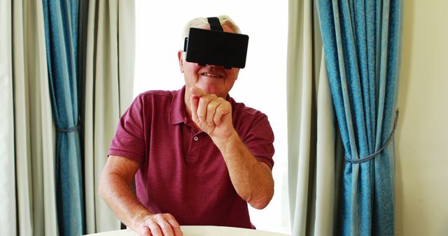 Elderly man sitting at a table, wearing a VR headset, enjoying immersive digital experience, indoors with blue curtains. Perfect for illustrating the adoption of latest technologies by senior citizens, elder care innovation, and promoting virtual reality products.