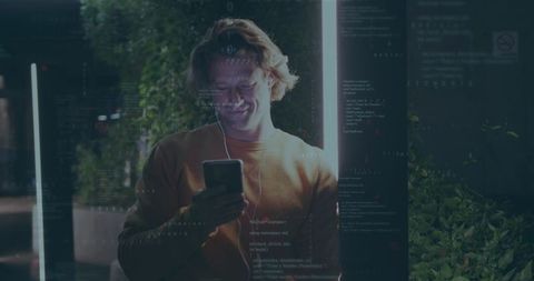 Young man listening on wired earbuds checking smartphone under neon lights with code overlay