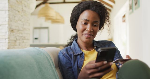 A woman is sitting comfortably on a couch at home, using a smartphone for online shopping. The atmosphere is relaxed and casual, creating a relatable and inviting scene. Ideal for promoting e-commerce, online payment services, technology for convenience, or articles about modern, digital lifestyles and consumer habits.