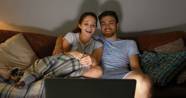 Young couple sitting on couch while watching a movie on laptop. They appear to be smiling and enjoying their time together. Suitable for content related to technology use in daily life, relationships, leisure activities, and home environment.