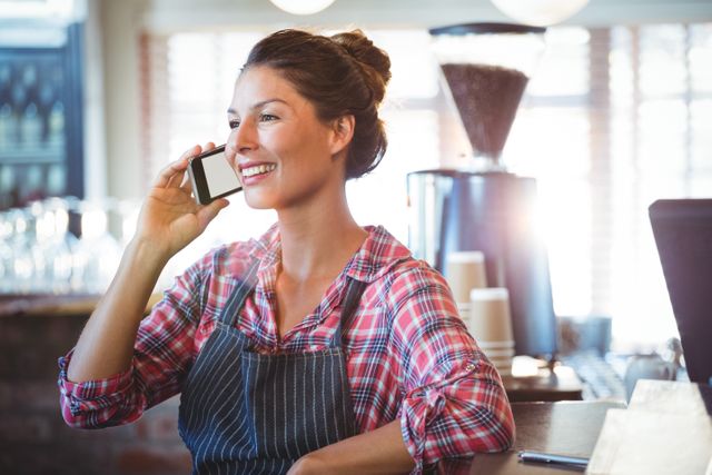 Waitress in a plaid shirt and apron smiling while making a phone call in a cozy cafe. Ideal for illustrating themes of customer service, hospitality, small business operations, and professional communication in a coffee shop or restaurant setting.