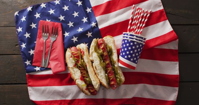 Hot dogs on an American flag with party supplies including napkins, forks, and cups. Ideal for representing patriotic celebrations like Fourth of July or Memorial Day. Suitable for use in articles, advertisements, and social media promoting national pride, summer picnics, and barbecues.