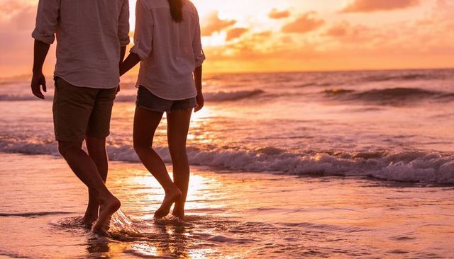 Romantic couple walking barefoot in ocean waves at sunset, golden hour stroll