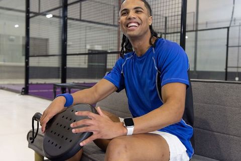 Energetic padel player smiling while resting on bench holding paddle in indoor court
