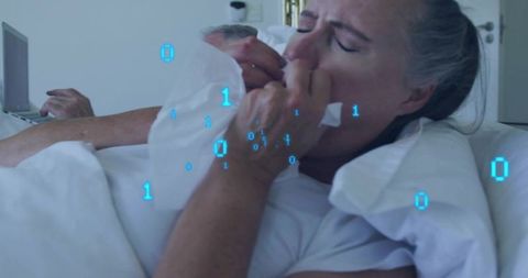 Mature woman pressing tissue to face in bed while partner using laptop with binary overlay