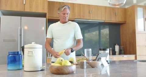 Mature man peeling banana at modern kitchen island with tablet and wooden bowl