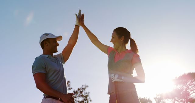 Couple celebrating success on golf course with high-five at sunset. Ideal for themes of teamwork, partnership, and outdoor activities. Perfect for use in promotional materials for golf courses, sports training, and healthy lifestyle campaigns.