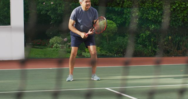 An elderly man intently playing tennis on a court, showcasing his focus and dedication to the sport. This image is perfect for illustrating active retirement lifestyles, senior fitness programs, and promotional materials for tennis training for older adults. It can also be used in health and wellness brochures, active aging campaigns, and community sports events aimed at seniors.