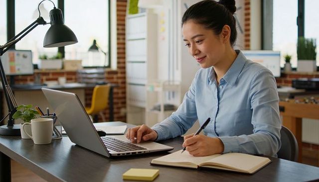 Asian professional woman working on laptop and taking notes in modern loft office
