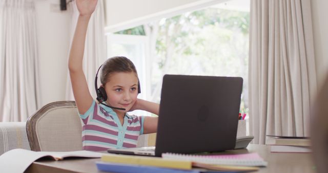 A young girl is actively participating in an online learning session from home, raising her hand while wearing a headset and working on a laptop. This visual can be used for illustrating remote education, children's e-learning experiences, and digital classroom settings. Ideal for educational blogs, school websites, e-learning platforms, and educational technology advertisements.