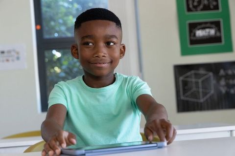 African american boy reaching for tablet at classroom desk, using technology to learn