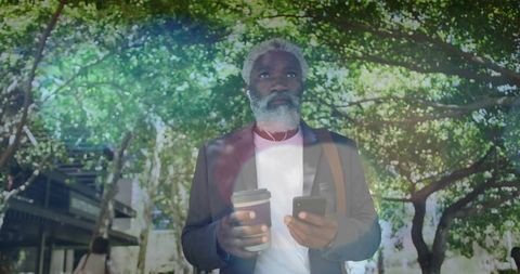 Midadult professional walking under trees checking smartphone while holding takeaway coffee