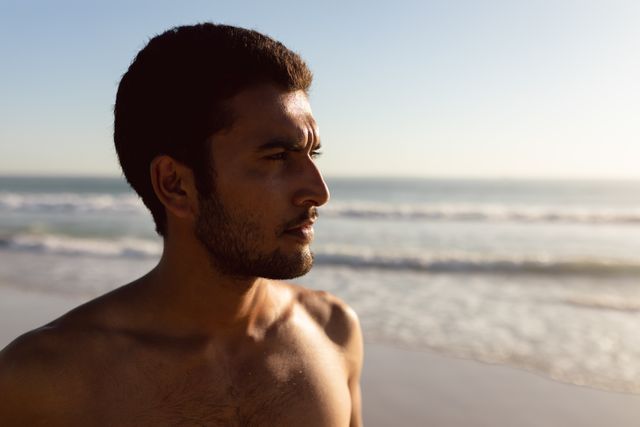 Young man standing on beach during sunset, looking thoughtful and serene. Ideal for use in lifestyle blogs, mental health articles, travel promotions, and advertisements focusing on relaxation and contemplation.