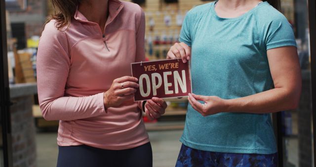 Two women holding an 'Open' sign, wearing casual activewear tops. This can be used for promoting businesses, especially fitness centers, gyms, or wellness centers. Perfect for highlighting reopening, welcoming new clients, or team spirit.