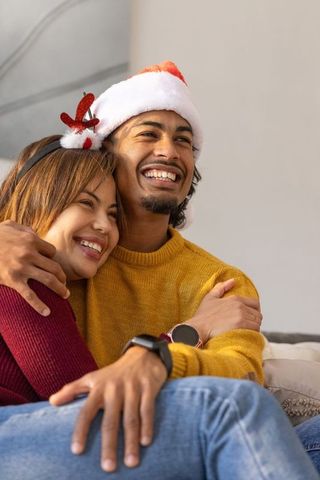 Smiling couple cuddling on sofa wearing santa hat and reindeer headband, smartwatch visible