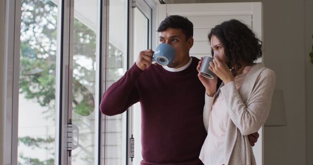 A couple is drinking coffee by a large window, enjoying a cozy and peaceful morning together. This image can be used to portray themes of relaxation, daily routines, love, togetherness, and domestic life. It is ideal for promotional materials on home comfort, lifestyle, or coffee products.