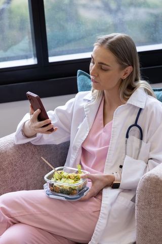 Female nurse taking lunch break checking phone while eating salad on sofa