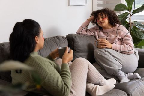 Diverse friends relaxing on sofa with mugs and under-eye gel patches, cozy chat