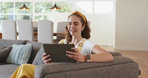 Young woman relaxing on modern gray sofa using tablet and smiling in bright living room
