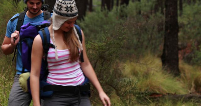 This image features a young couple hiking in a forest, wearing winter hats and carrying backpacks. It evokes themes of outdoor adventure, nature exploration, physical activity, relaxation, and bonding. Ideal for use in travel blogs, advertisements for outdoor gear, nature magazines, and lifestyle promotions related to health and fitness.
