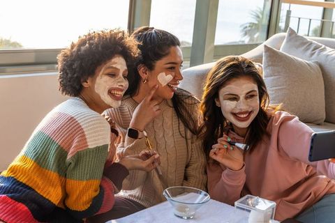 Diverse friends applying face masks and taking selfie on sunlit living room sofa