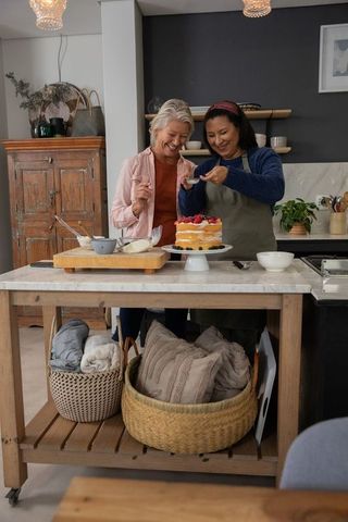 Senior women decorating layered berry cake on kitchen island wearing aprons and smiling