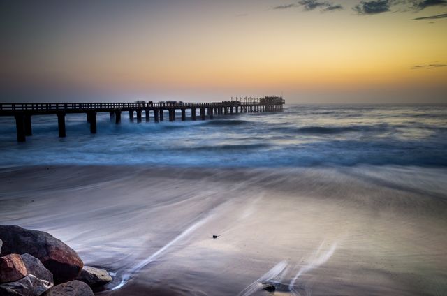 This peaceful beach scene at sunset shows a long pier extending into the ocean under a colorful sky. Gentle waves lap at the shore, creating a tranquil mood perfect for themes like relaxation, travel, vacation destinations, and nature's beauty. Great for websites, travel brochures, and peaceful mood illustrations.