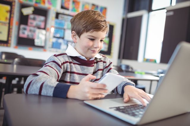 Smiling schoolboy using laptop and mobile phone on desk at classroom