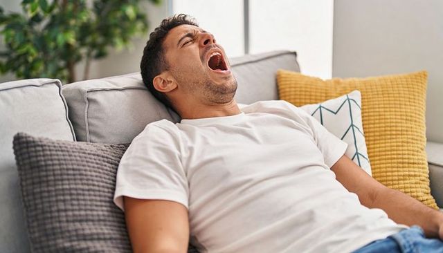Yawning hispanic man reclining on gray sofa, relaxing at home in white tee and blue jeans