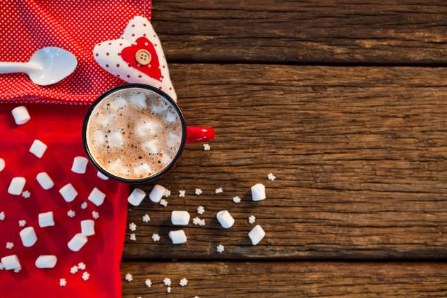 Hot chocolate with marshmallows in a red mug on a rustic wooden table. A red napkin and heart decoration add a festive touch, making it perfect for Christmas or winter-themed content. Ideal for holiday promotions, cozy winter scenes, or festive social media posts.