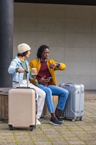 Multicultural travelers waiting at station checking watch and phone with carry-on luggage