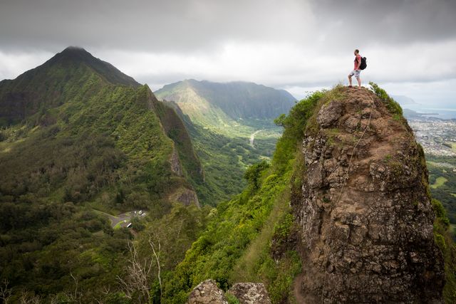 A lone hiker stands on the edge of a rocky cliff, admiring the expansive green mountain ranges and valleys below. This image captures the spirit of adventure and exploration, ideal for use in travel blogs, adventure tourism promotions, outdoor activity advertisements, motivational posters, and nature magazines.