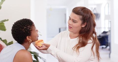Diverse friends sharing skincare routine, applying moisturizer together in bright living room