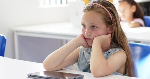 Pensive schoolgirl resting chin on hands with tablet on desk in bright classroom