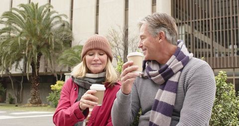 Senior couple enjoying takeaway coffee on urban bench wearing scarves smiling on autumn morning