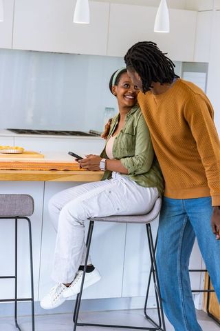African american couple smiling and leaning at modern kitchen breakfast bar holding smartphone