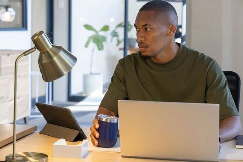 African american man working at modern desk holding blue tumbler and using laptop