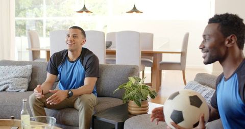 Diverse male teammates watching soccer and chatting on gray sofa at home with soccer ball