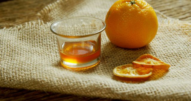The image shows a glass containing a small amount of orange juice next to a whole orange and several slices of dried orange on a burlap fabric. This image can be used for promoting healthy eating, organic products, or fresh fruit beverages. It conveys a natural and rustic feel, making it suitable for health blogs, recipe websites, and organic produce advertisements.